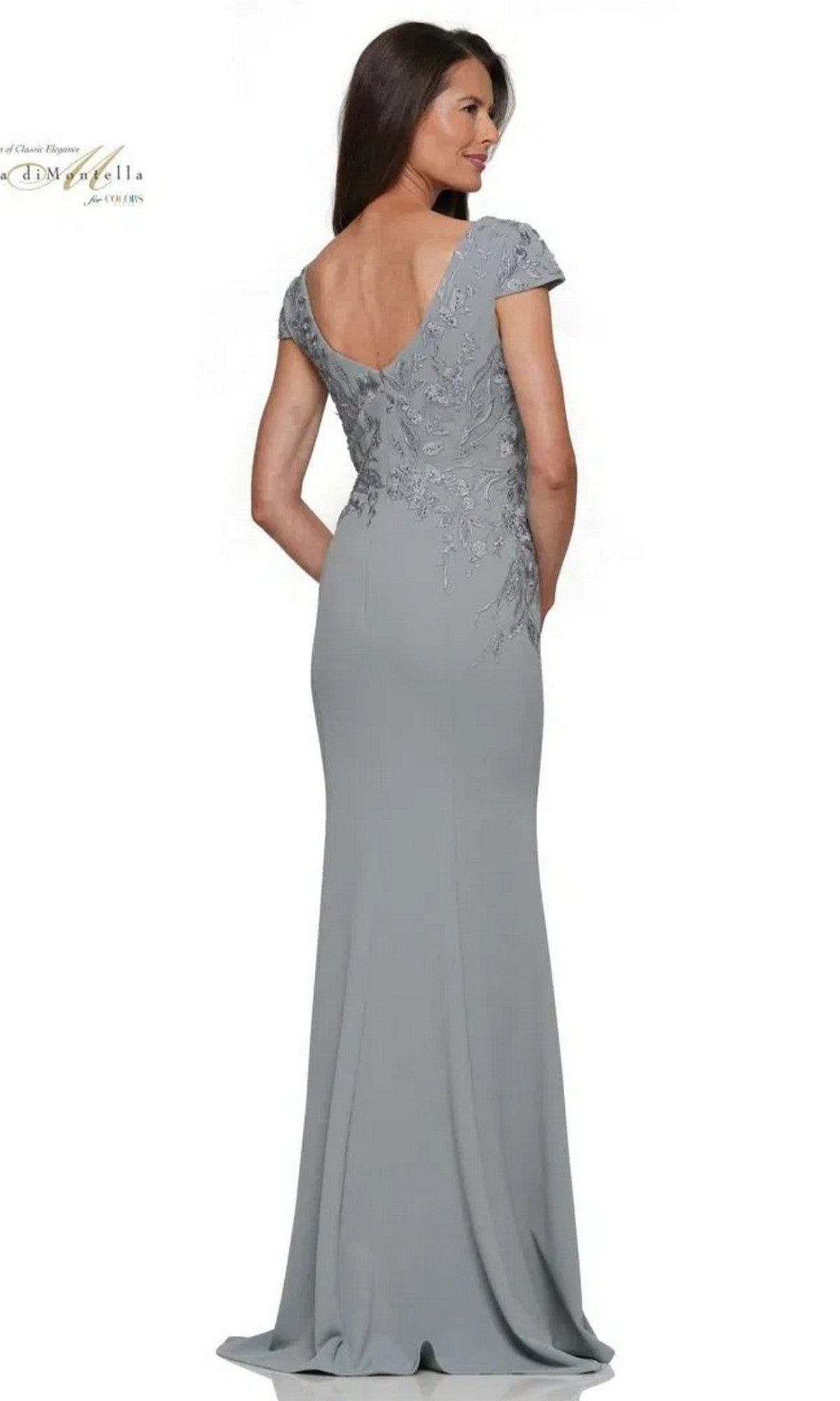 Woman wearing the Rina Di Montella RD2960, a floor-length light gray formal dress with an embroidered bodice and short sleeves, stands with her back to the camera and hands in pockets.